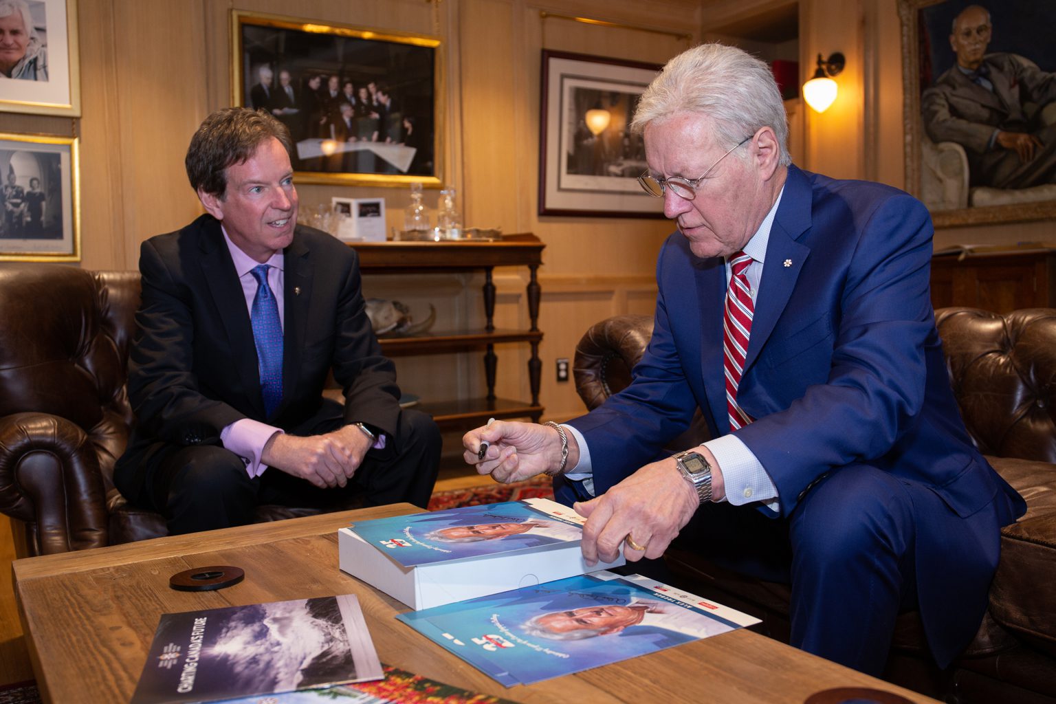 Alex Trebek signs posters while seated across from John Geiger