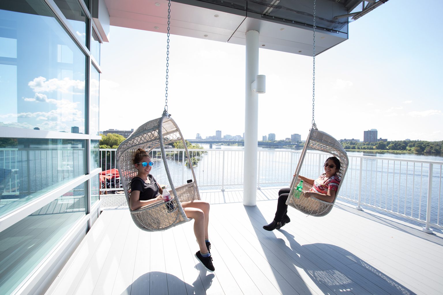 Teenage girls sit in two wicker swing chairs suspended from the roof of a balcony overlooking a river