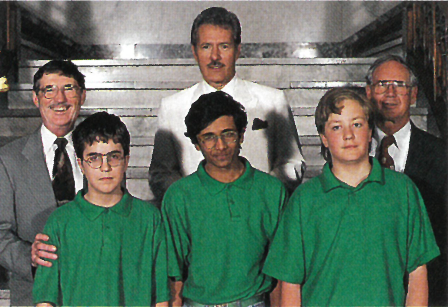 Three men in suits stand behind three young men in green polo shirts
