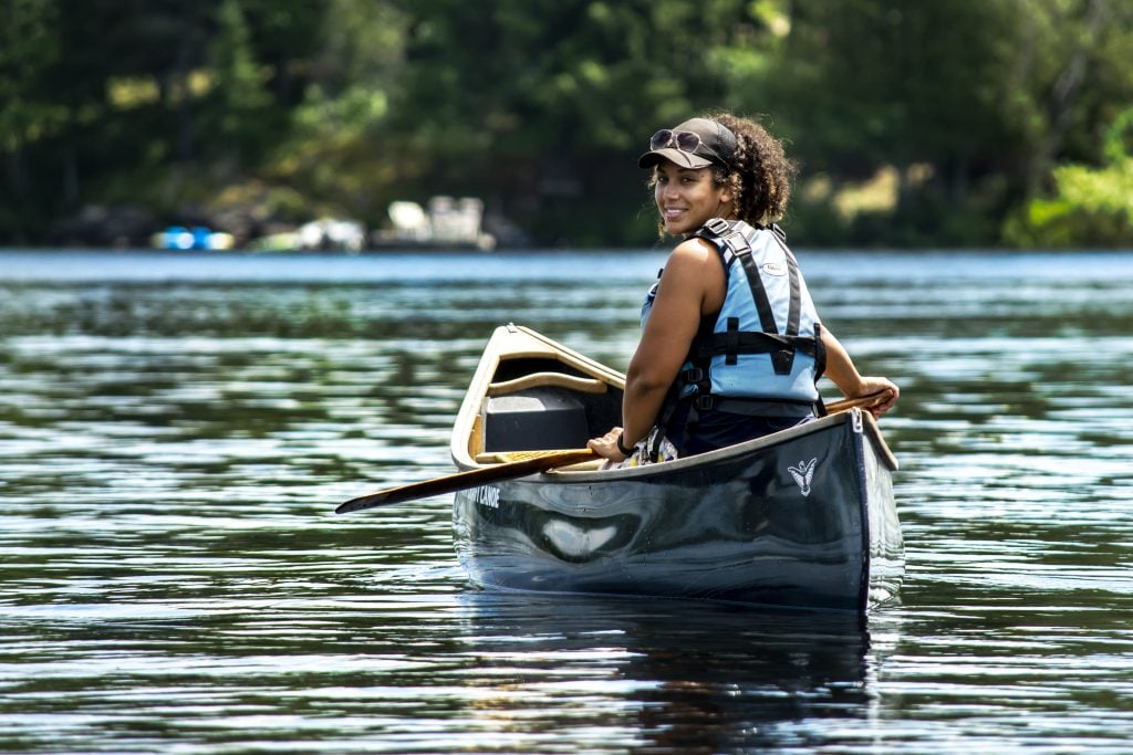 Tori, in her canoe, smiles back towards the camera across glistening blue water