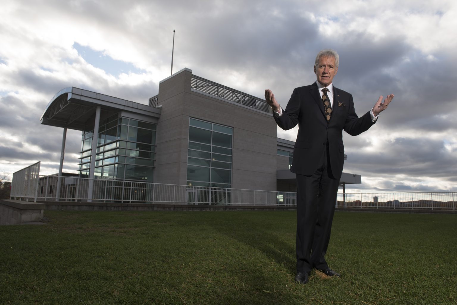 Alex Trebek stands on grass with the Royal Canadian Geographical Society Headquarters behind him