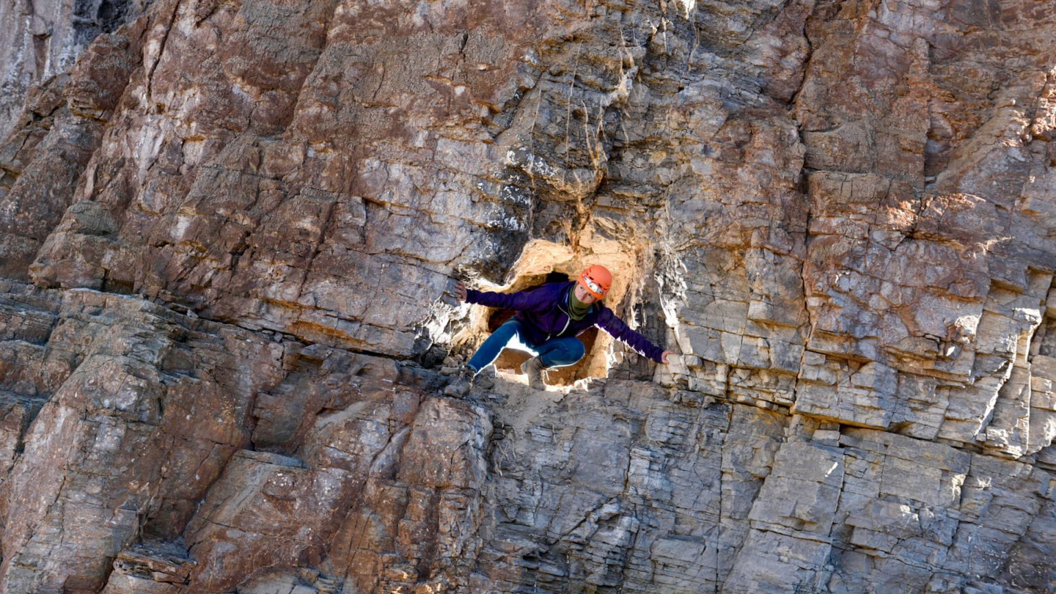 Gina Moseley, a white woman wearing a purple jacket and an orange hard hat, grouches in an opening in a rust-orange cliff face.