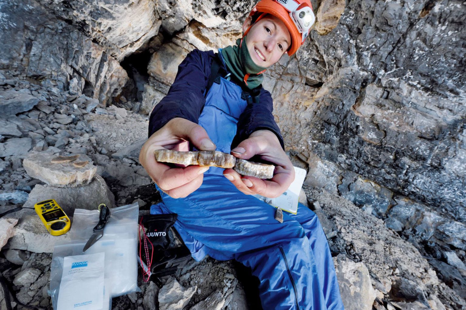 Gina Mosely, a white woman wearing blue overalls and an orange hard hat, shows a piece of mineral deposit to the camera