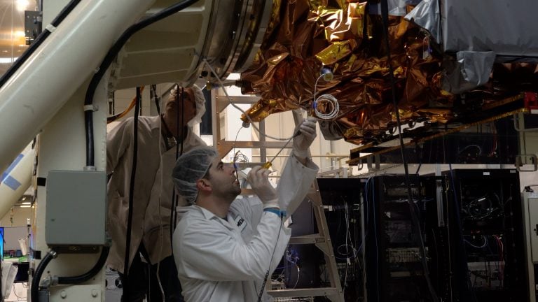 A worker in a flightsuit makes adjustments to a satellite