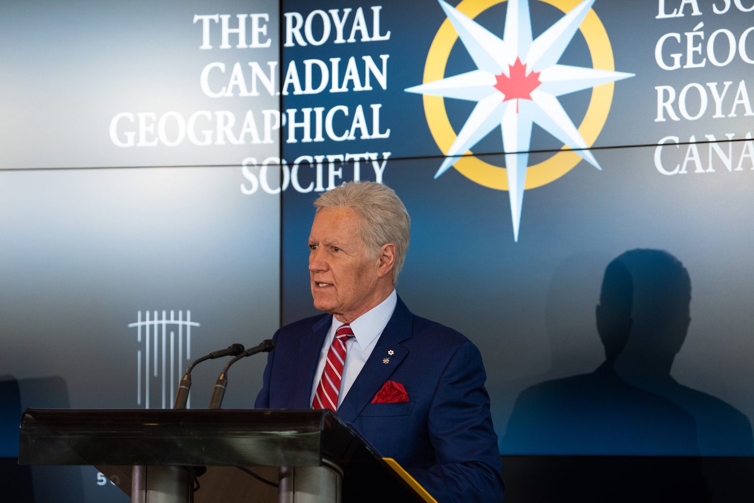 Alex Trebek stands at a podium with the Royal Canadian Geographical Society logo on the screen behind him