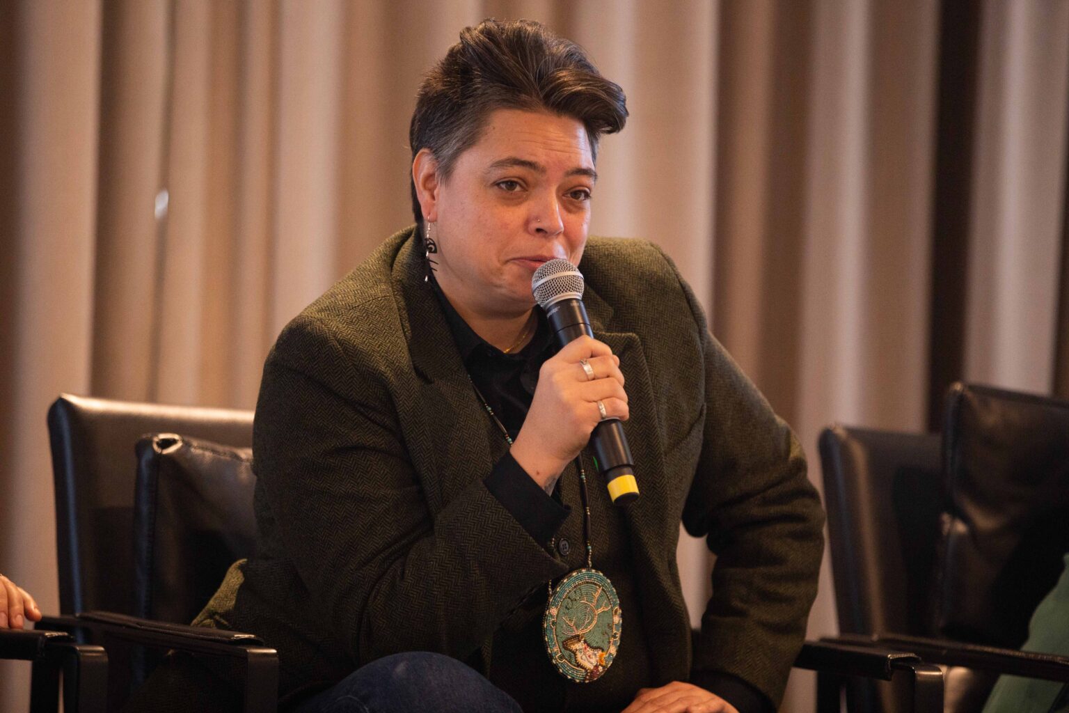 Valerie Courtois, an Innu woman, sits and speaks into a microphone. She wears a serious expression on her face and a beaded medallion depicting a caribou