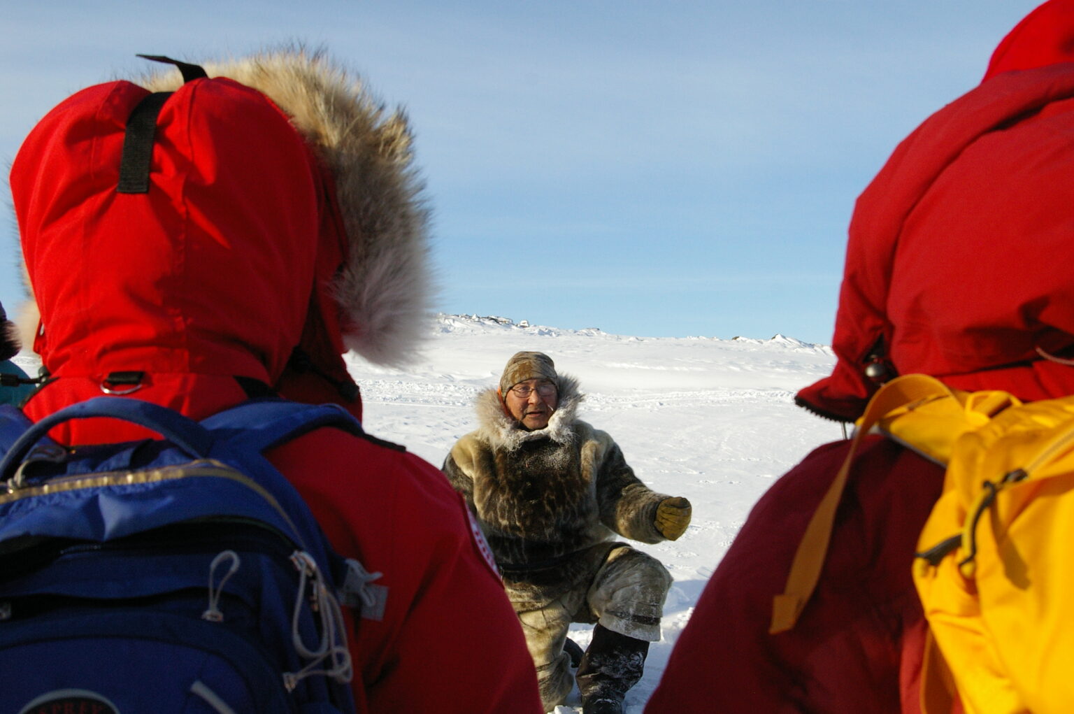 An Inuit man wearing furs speaks to a group of students who are wearing red jackets with their hoods up. It is a sunny day and the ground is snow.
