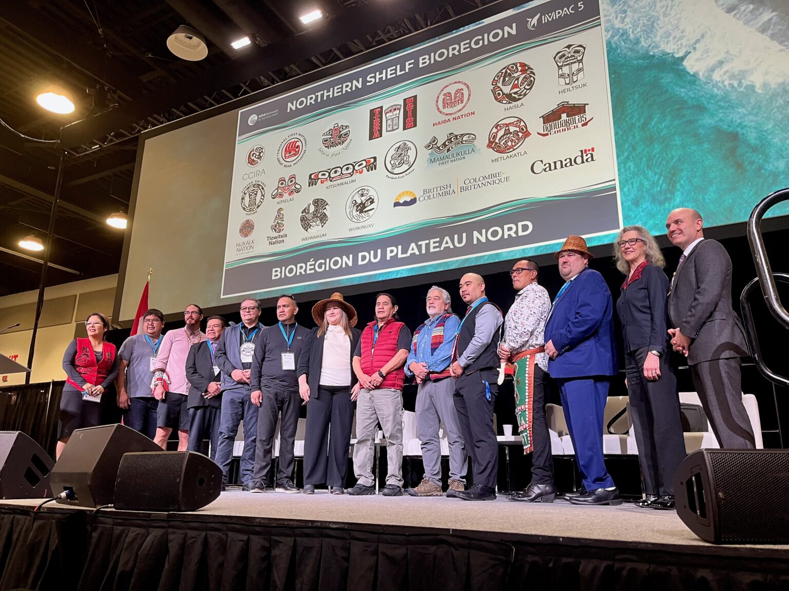 14 people stand on a stage with a screen behind them showing the logos of 15 first nations, as well as the British Columbia and Canada governments.