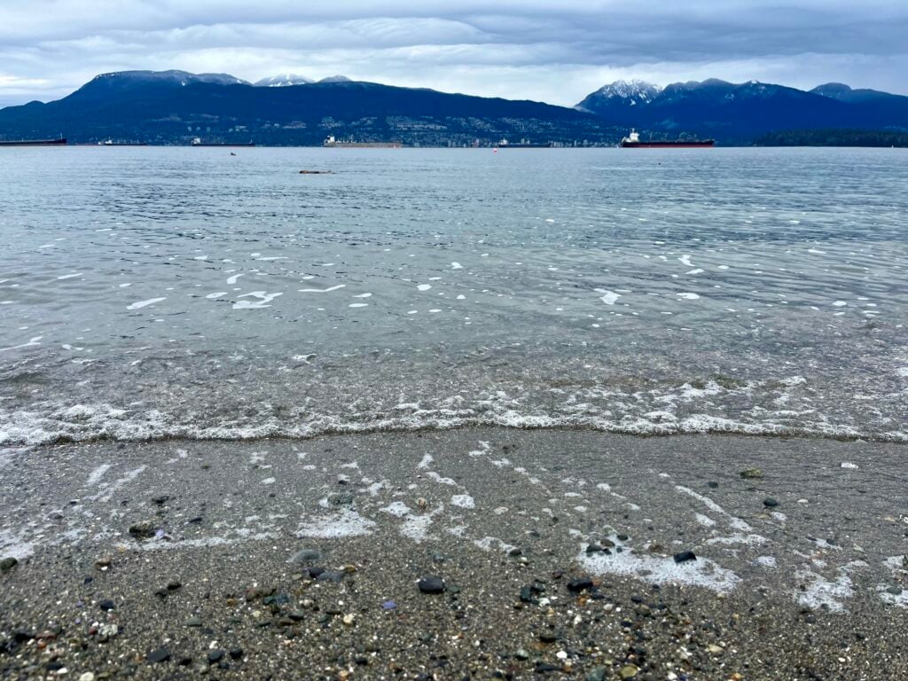 Gentle waves lap onto the shores of a pebbly beach as blueish mountains loom in the background.