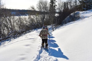 A group of people snowshoe down a snowy bank beside a frozen river, their shadows stretching out over sparkling snow. There is a bridge in the background and the snow is low in the sky. At the back is a woman wearing a knitted fleece and a Métis scarf tied around her waist.