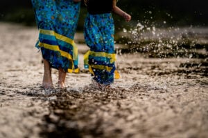 Two woman wearing ribbon skirts wade in the shallows, splashing water into a golden spray around them