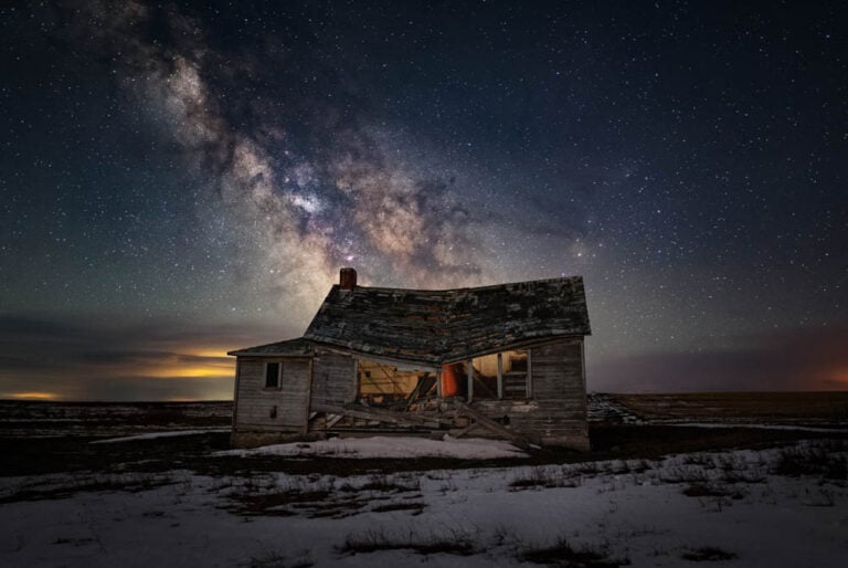 the milkway arcs in a blue and orange sky above a gently-lit abandoned building
