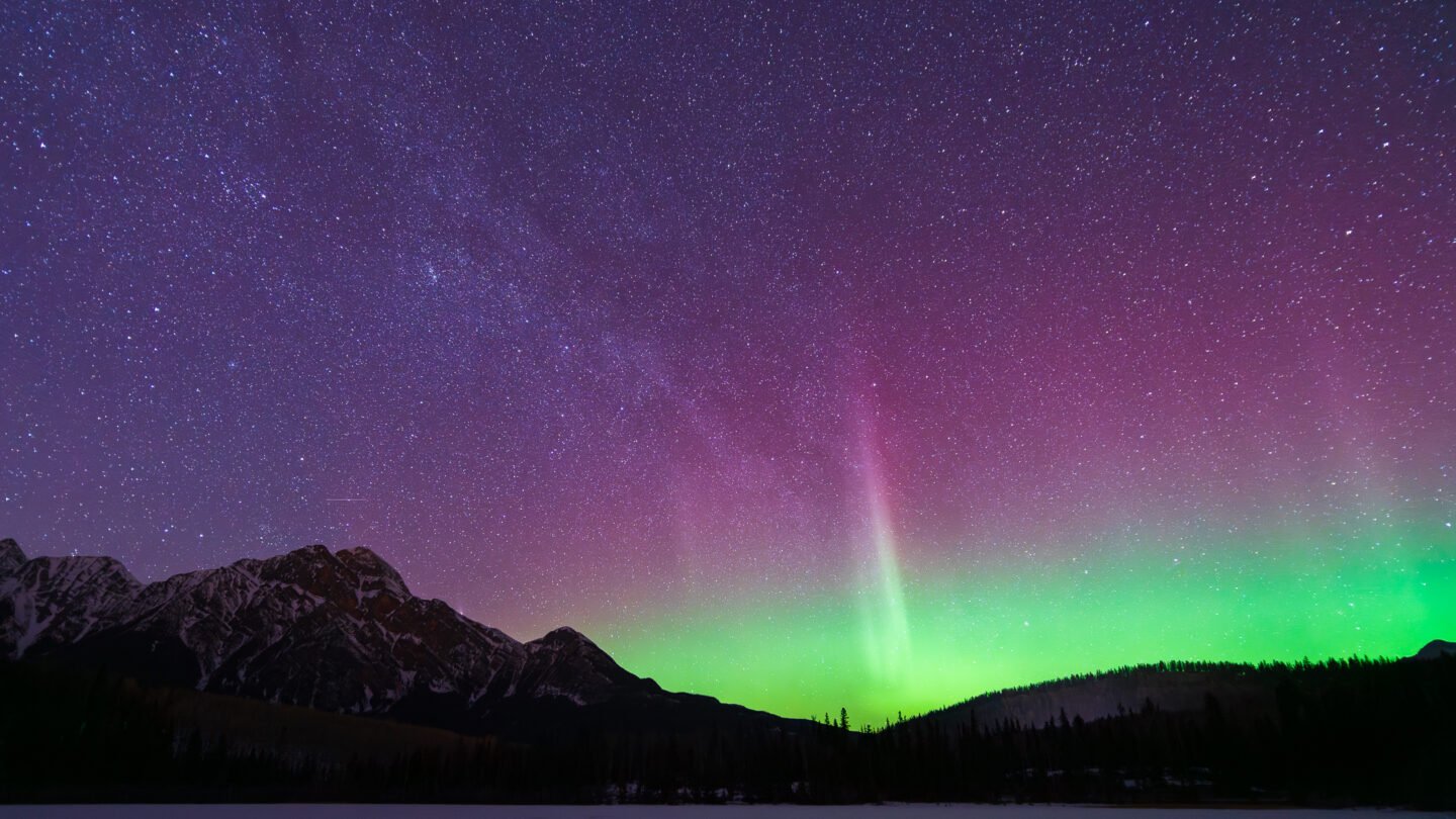 green aurora shoots up into a purple and violet sky spangled with stars. There are silhouetted mountains in the foreground.