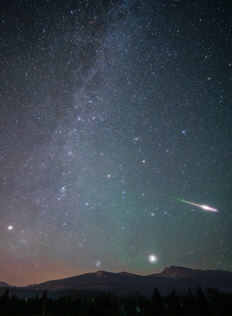 Above a mountain silhouette, the sky is full of many stars and the shape of the milky way. A meteor can be seen streaking through the sky.