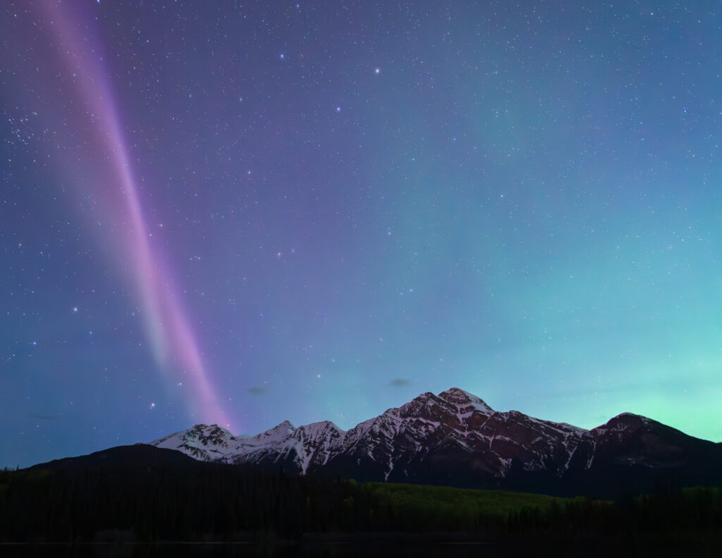 a pink thread of light winds through a glowing blue and green sky. There are mountains silhouetted in the foreground.