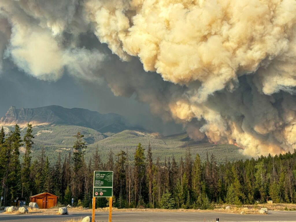 brown smoke balloons into the sky above a forested mountain. In the foreground is a deserted parking lot of a trailhead.
