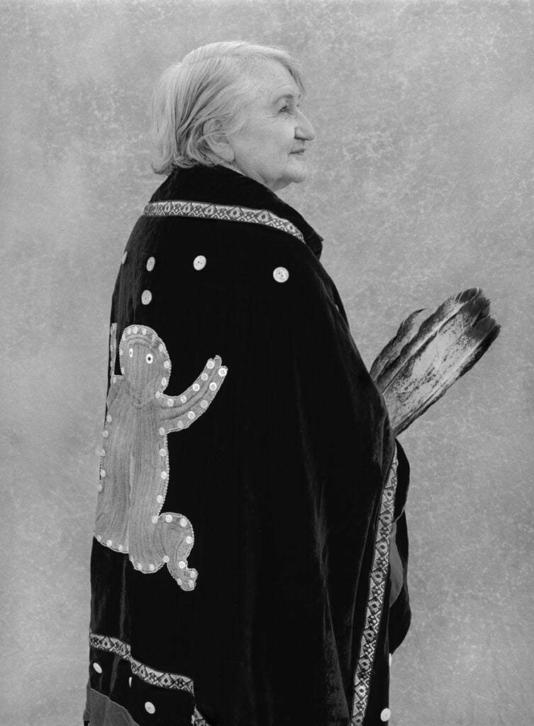 a black and white portrait of Maggie Hodgson holding feathers and wearing a shell blanket of the Frog Clan of the Carrier peoples from the northern interior of B.C.