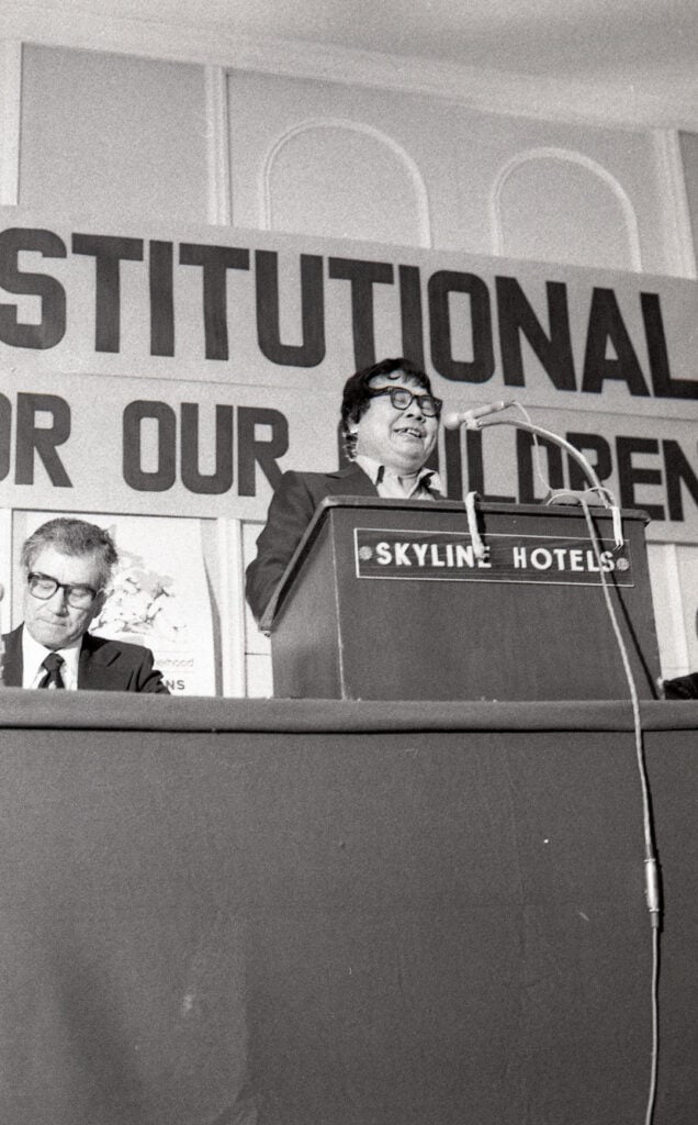 a man stands at a podium. behind him, part of a sign reads 