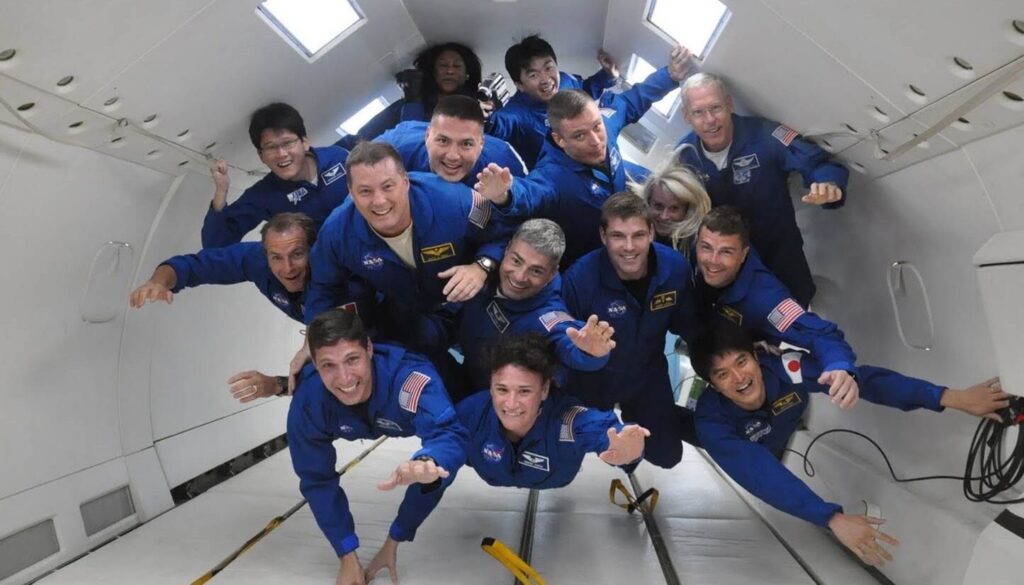 A group of 15 astronauts wearing blue flight uniforms float around in a white room.