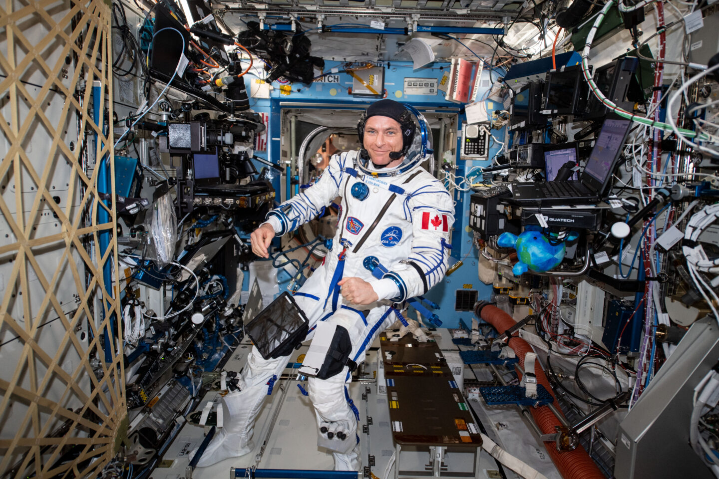 Astronaut David Saint-Jacques in a white pressure suit surrounded by a multitude of wires and space equipment.
