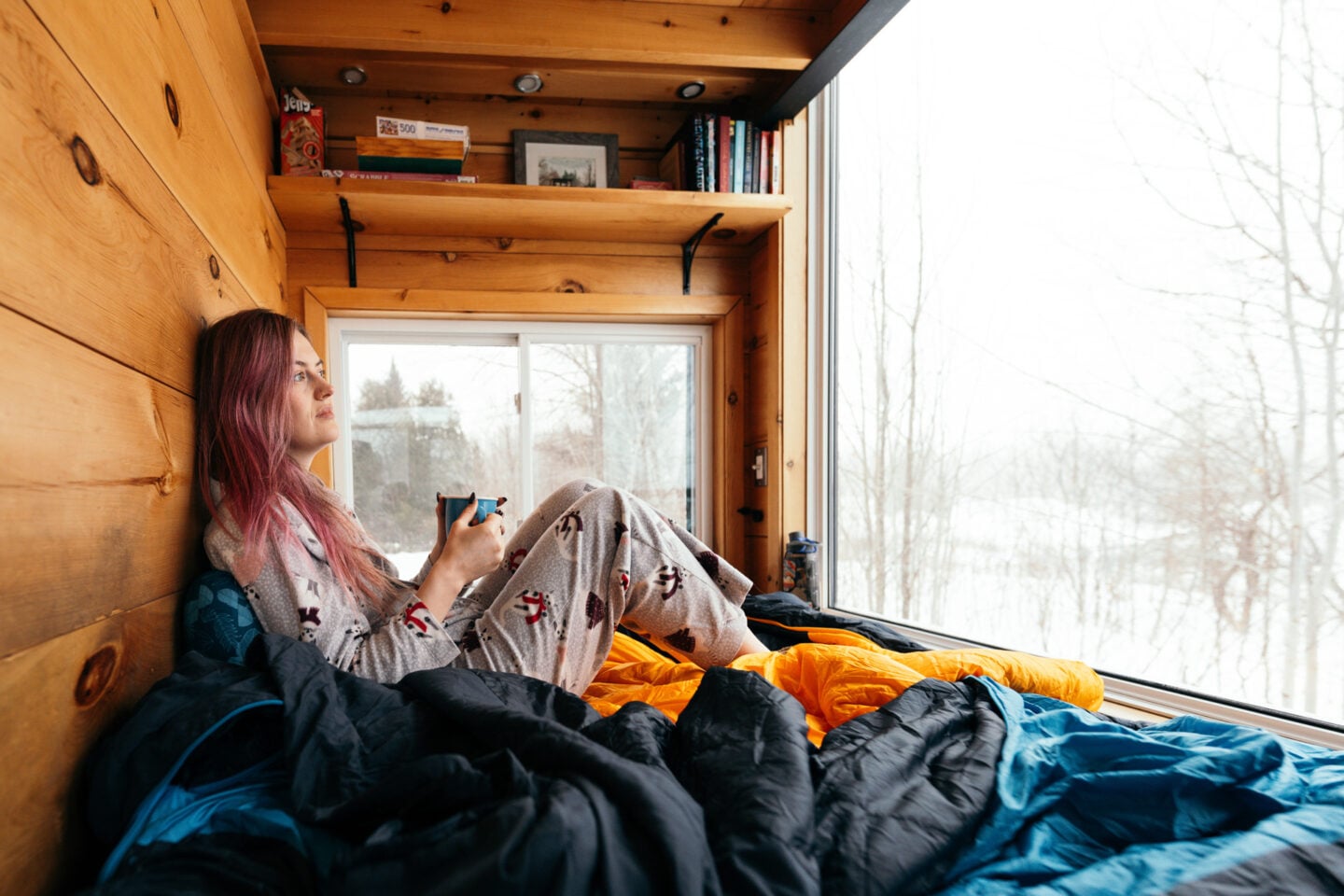 a white woman with pink hair sits on a sleeping bag looking out of the window while drinking a cup of tea