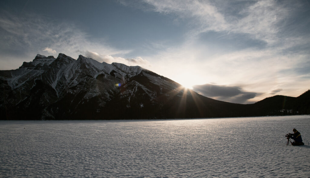 A man kneels behind a camera on a frozen landscape with a large mountain in the background.