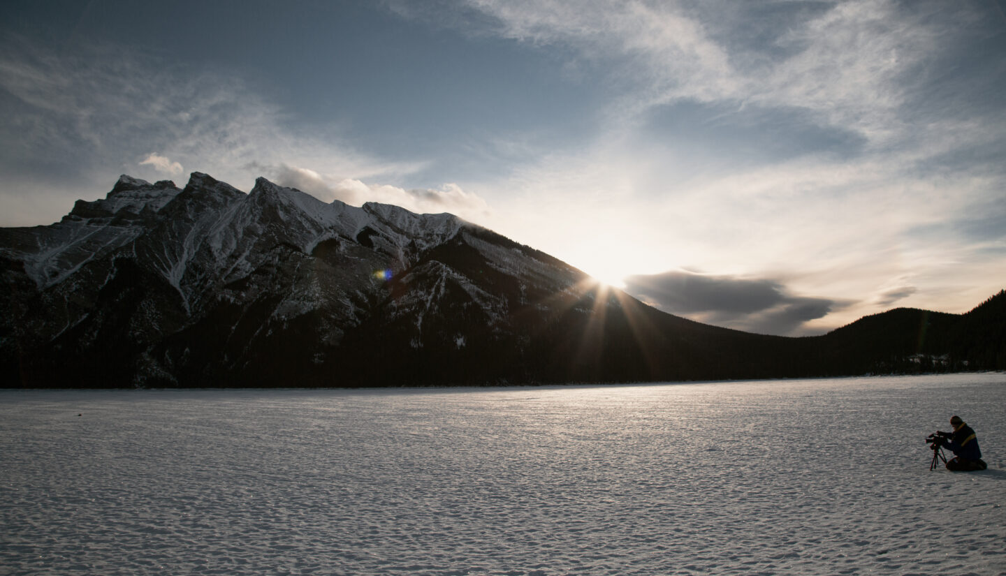 A man kneels behind a camera on a frozen landscape with a large mountain in the background.