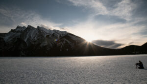 A man kneels behind a camera on a frozen landscape with a large mountain in the background.