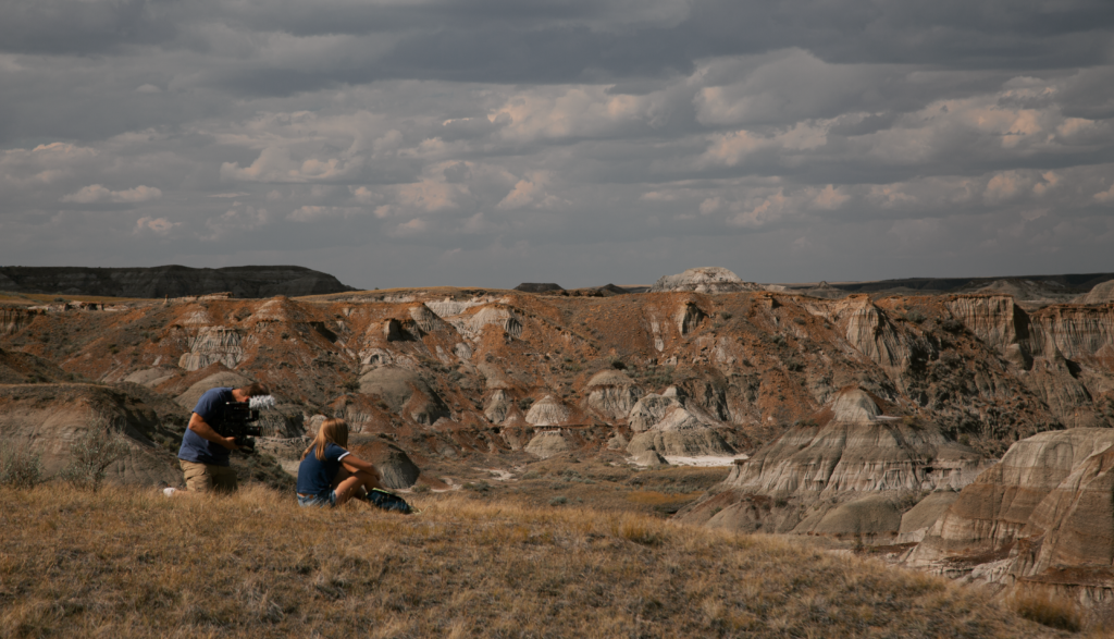 A man films a girl staring into the distance while surrounded by a dry landscape.