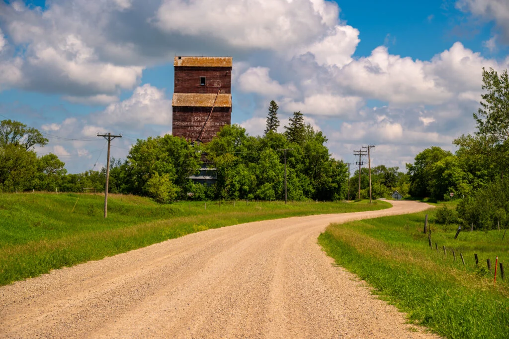 Historic wooden grain elevator overlooking a winding gravel road in Lenore, Manitoba beneath a blue sky with large clouds.