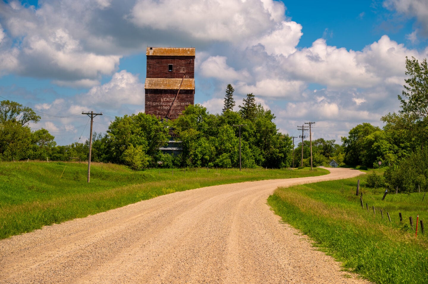 Historic wooden grain elevator overlooking a winding gravel road in Lenore, Manitoba beneath a blue sky with large clouds.